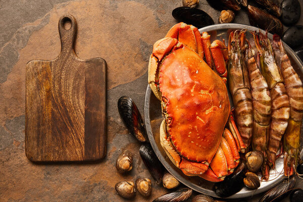 top view of uncooked crab, shellfish, cockles and mussels in bowl near wooden chopping board on textured surface 