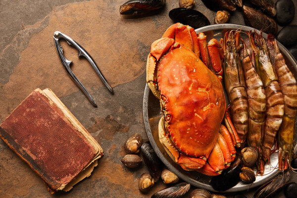 top view of uncooked crab, shellfish, cockles and mussels in bowl near seafood cracker and crumbly old book on textured surface 