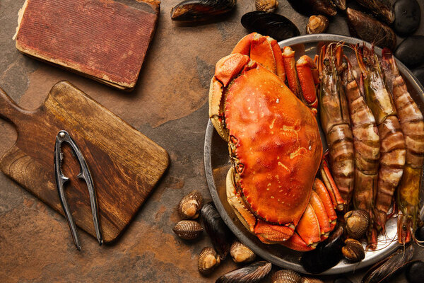 top view of uncooked crab, shellfish, cockles and mussels in bowl near crumbly old book and seafood cracker on wooden chopping board on textured surface 