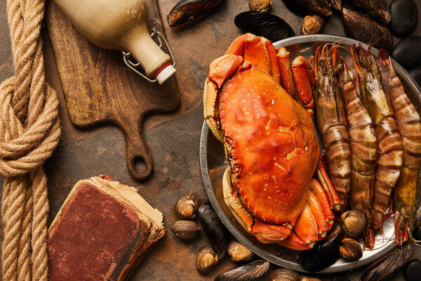 top view of uncooked crab, shellfish, cockles and mussels in bowl near crumbly old book, rope and bottle with cork on wooden chopping board on textured surface 