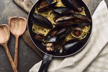 top view of delicious pasta with mollusks and mussels in frying pan on wooden cutting board near napkin and spatulas clipart