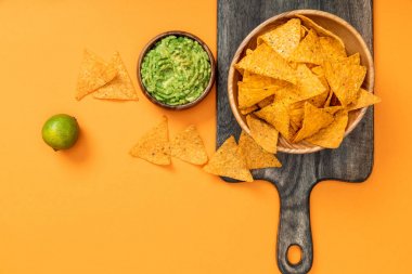 ahşap kesme tahtası üzerinde gevrek Meksika nachos üst görünümü, turuncu arka plan üzerinde guacamole ve kireç