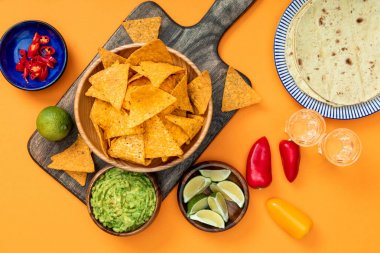 top view of crispy Mexican nachos served on wooden cutting board with guacamole, peppers, limes, Tequila and tortillas on orange background clipart