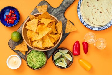 Mexican nachos served on wooden cutting board with guacamole, cheese sauce, peppers, limes, Tequila and tortillas on orange background clipart