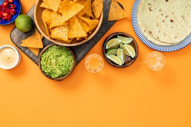 Mexican nachos served on wooden cutting board with guacamole, cheese sauce, peppers, limes, Tequila and tortillas on orange background with copy space clipart
