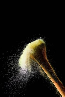 cosmetic brush with yellow colorful scattered powder on black background clipart
