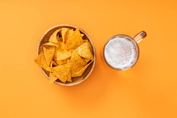 top view of crispy nachos in wooden bowl near glass of beer on orange background, Mexican cuisine