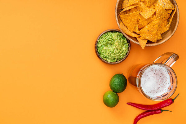 top view of crispy nachos, guacamole, beer, limes and chili peppers on orange background with copy space