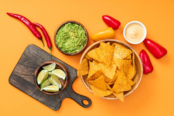 top view of crispy Mexican nachos, guacamole, peppers, limes, cheese sauce and wooden cutting board on orange background
