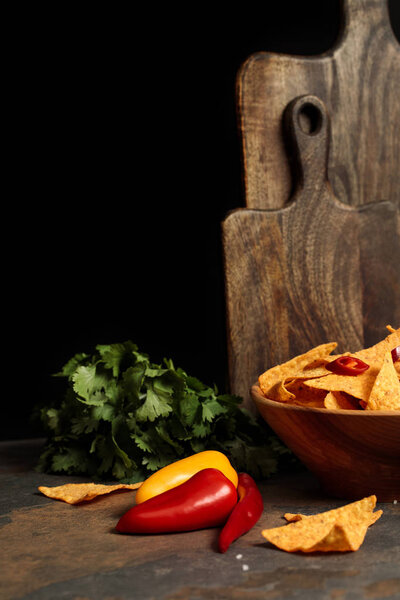 Mexican nachos with salt and chili peppers near wooden cutting boards and parsley on stone table isolated on black