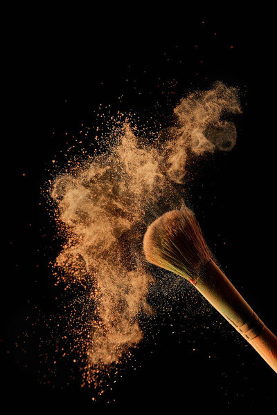 косметическая кисть с красочным оранжевым порошковым облаком на черном фоне
