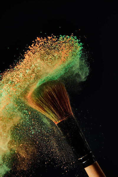 косметическая кисть с зеленым и оранжевым взрывом порошка на черном фоне

