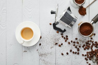 Top view of  cup of coffee on saucer near geyser coffee maker, portafilter and spoon on white wooden surface with coffee beans clipart