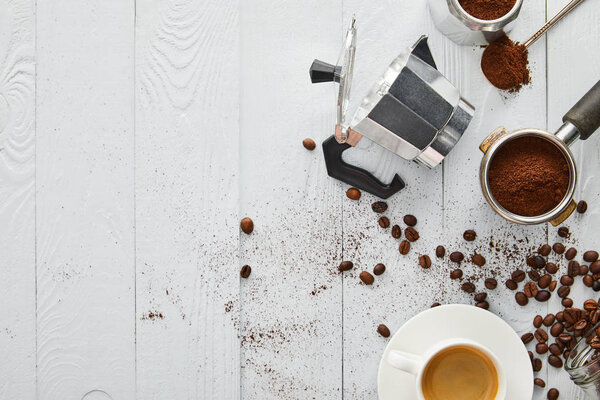 Top view of geyser coffee maker near portafilter, spoon and cup of coffee on white wooden surface with coffee beans