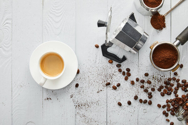 Top view of  cup of coffee on saucer near geyser coffee maker, portafilter and spoon on white wooden surface with coffee beans