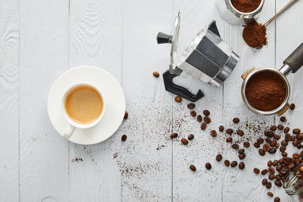 Top view of cup of coffee on saucer near geyser coffee maker, portfilter and spoon on white wooden surface with coffee beans
