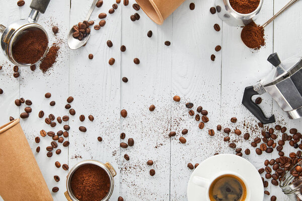 Top view of portafilters, spoons, paper cups, geyser coffee maker and cup of coffee on white wooden surface 