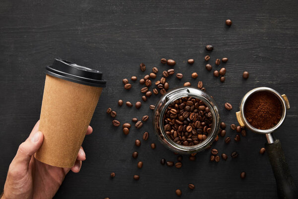 Top view of hand with paper cup under dark wooden surface with glass jar and portafilter