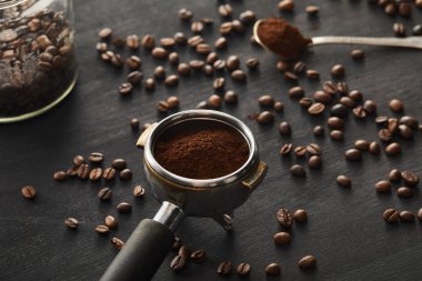 Portafilter filled with ground coffee near spoon ang glass jar on dark wooden surface with coffee beans clipart