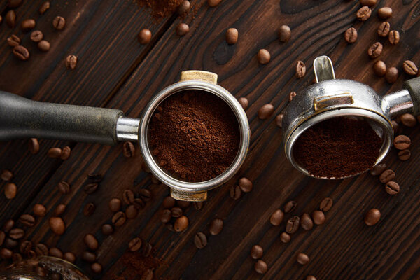Portafilters with ground coffee on dark brown wooden surface with coffee grains