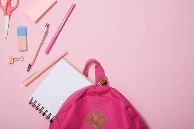 top view of scattered school supplies and blank notebook near backpack isolated on pink clipart