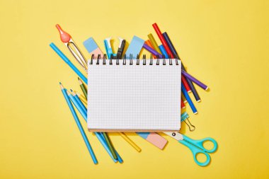 top view of colored pencils, scissors, rubbers and felt pens scattered near blank notebook on yellow clipart