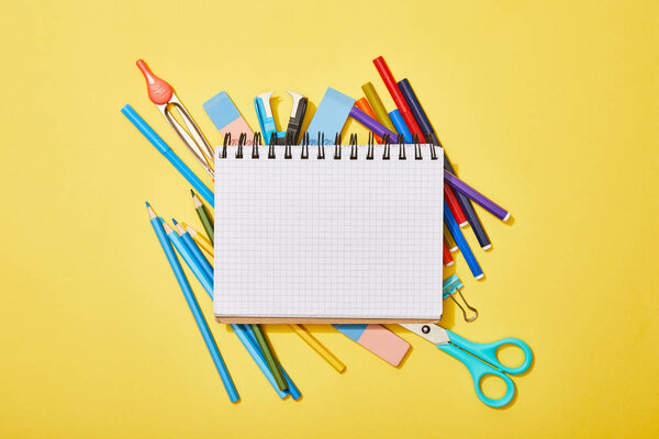 top view of colored pencils, scissors, rubbers and felt pens scattered near blank notebook on yellow