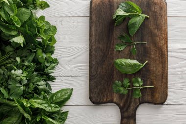 Top view of brown wooden cutting board with parsley, basil, cilantro and peppermint leaves near bundles of greenery on white surface clipart