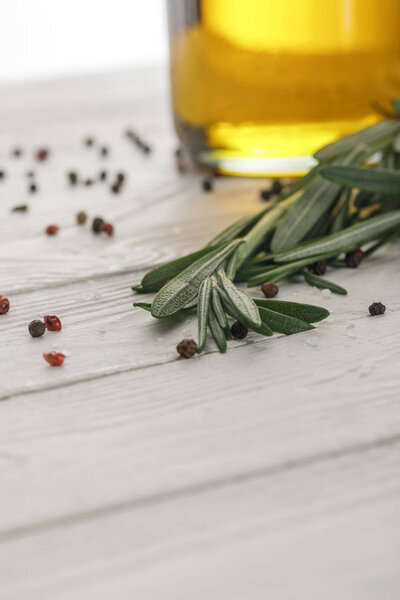Rosemary bundle near bottle with oil and scattered red and black peppercorns on white wooden surface 