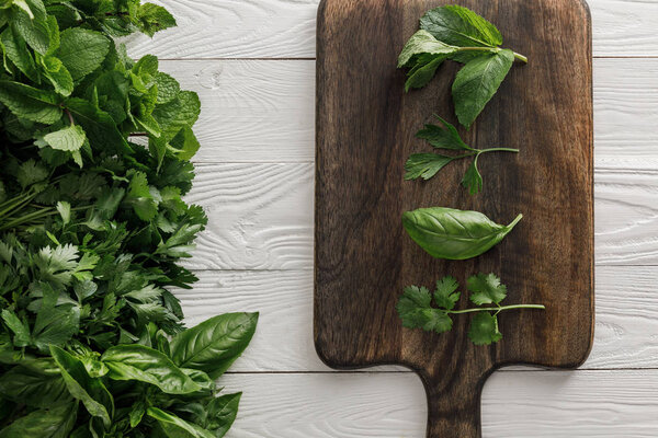 Top view of brown wooden cutting board with parsley, basil, cilantro and peppermint leaves near bundles of greenery on white surface