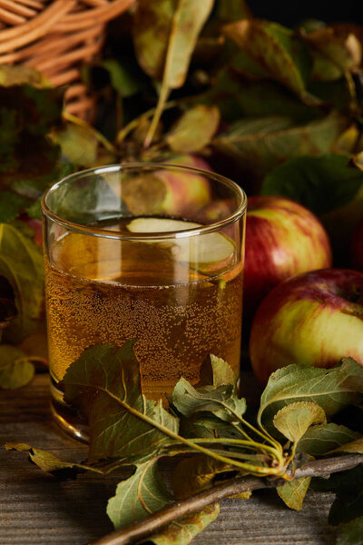 glass of cider near wicker basket and apples on wooden surface