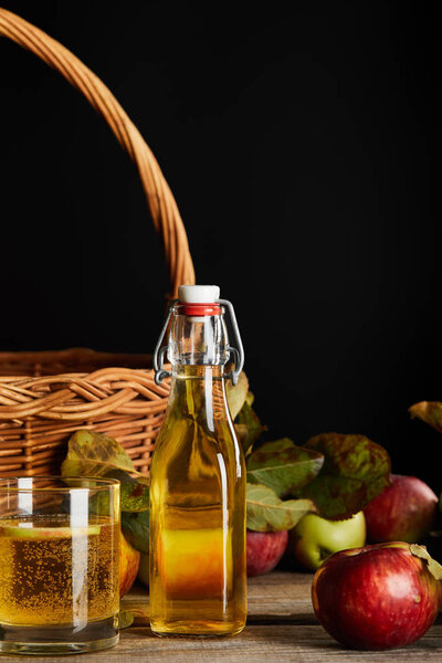 bottle and glass of cider near wicker basket and apples on wooden surface isolated on black
