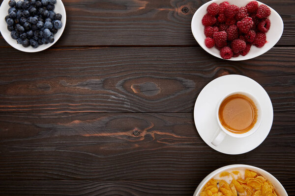 top view of bowl with cornflakes near cup of coffee and plates of raspberry and blueberry on wooden surface