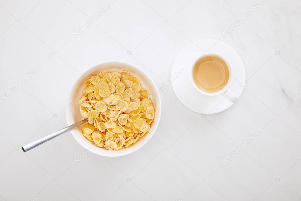 top view of bowl of cornflakes with spoon near cup of coffee on white marble surface