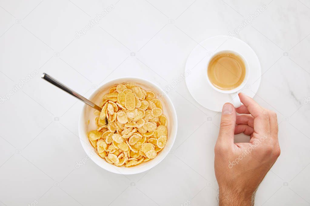 cropped image of man holding cup of coffee near bowl with cornflakes on white marble background
