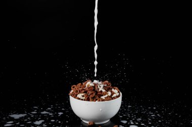 white bowl with assorted cereal with milk stream isolated on black clipart