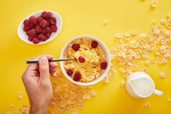 裁剪视图的人拿着勺子在碗与玉米片附近的新鲜树莓和牛奶壶隔离在黄色 — 图库照片
