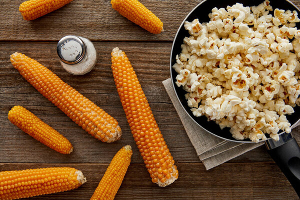 вид вкусного попкорна на сковородке рядом с кукурузой и солью на деревянном фоне
