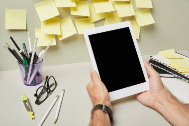 Kırtasiyenin yanında boş ekran ve gri ve beyaz üzerine not defterleri olan dijital tablet tutan adamın kırpılmış görüntüsü