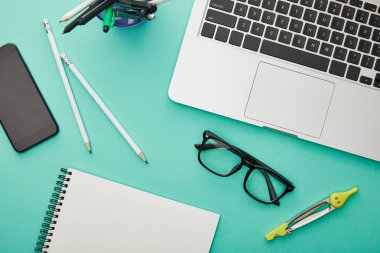 top view of gadgets near glasses, stationery and notebook isolated on turquoise clipart