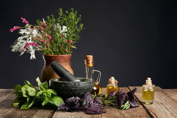 grey mortar near clay vase with herbs and small bottles on wooden table isolated on black
