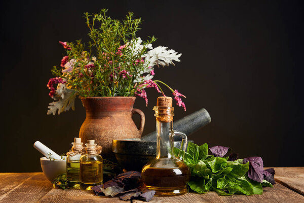 clay vase with fresh herbs and flowers near mortar and pestle and bottles on wooden table isolated on black