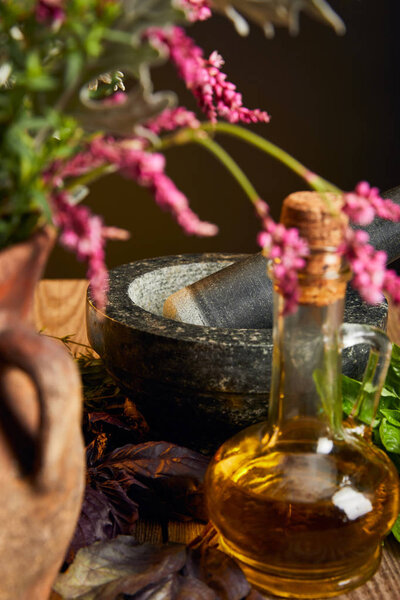 mortar with pestle near bottle with oil and vase with fresh wildflowers on wooden surface isolated on black