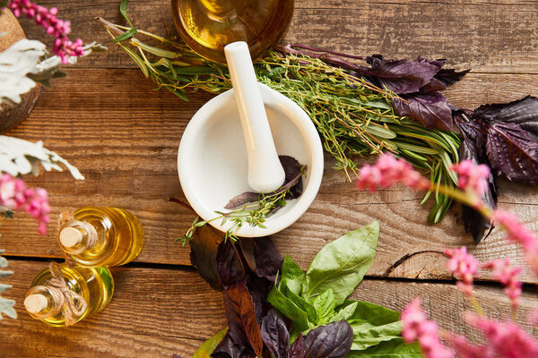 top view of mortar with pestle near bottle and fresh leaves and flowers on wooden surface