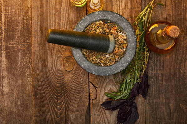 top view of mortar with pestle near fresh herbs and bottles on wooden surface with copy space