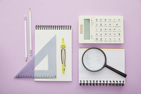 top view of magnifier on notebook near calculator and stationery isolated on purple 