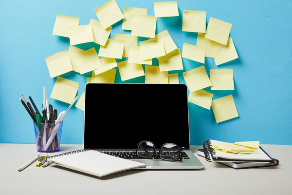 ноутбук с чистым экраном рядом с липкими нотами, ноутбуками и канцелярскими принадлежностями на сине-белом
 