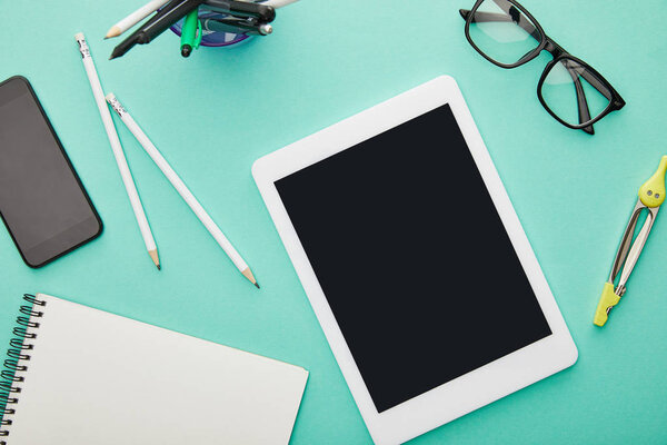 вид сверху на гаджеты с пустыми экранами возле очков, канцелярских принадлежностей и ноутбука, изолированных на бирюзовом
