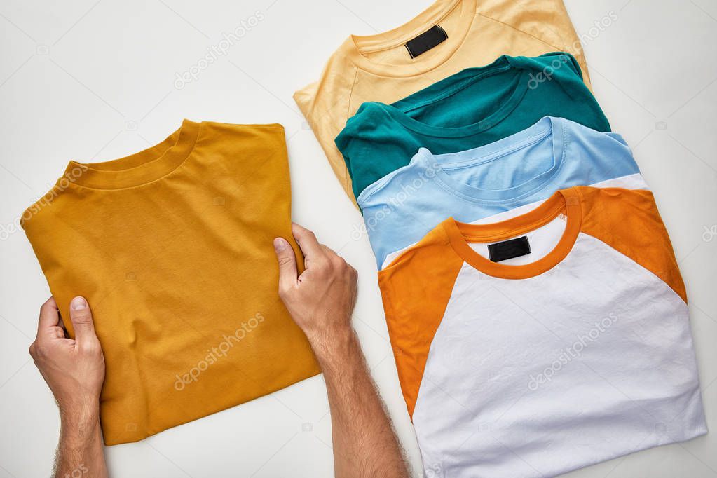 Cropped view of man holding ochre t-short near beige, orange, turquoise and blue ones on white background
