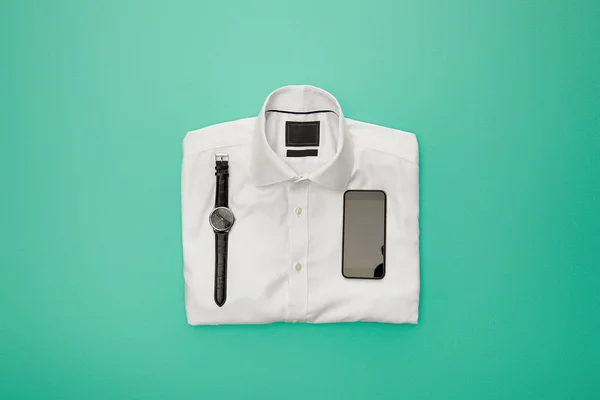手表和智能手机的顶视图 在绿松石背景上的普通白色折叠衬衫 — 图库照片